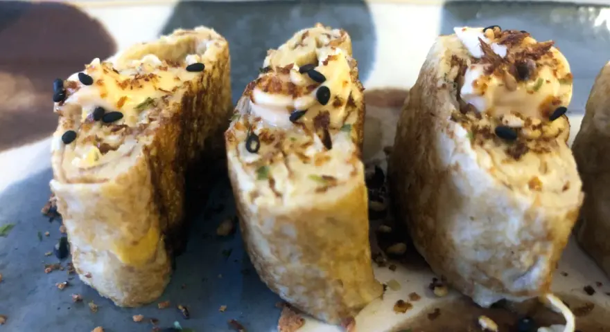 Trzy kawałki sushi w omlecie, polane majonezem i posypane czarnym sezamem.