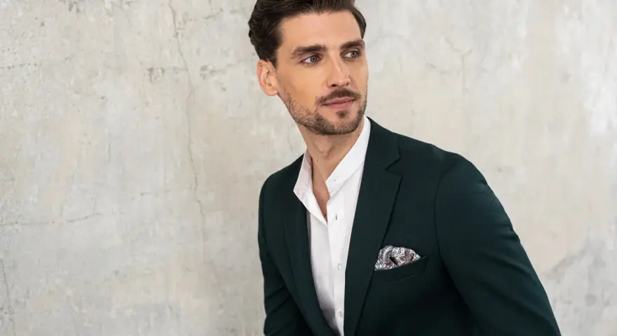 Elegancki mężczyzna w ciemnozielonej marynarce i białej koszuli z ozdobną poszetką. Jego kołnierzyk koszuli, czyli stójka, jest lekko rozpięty.