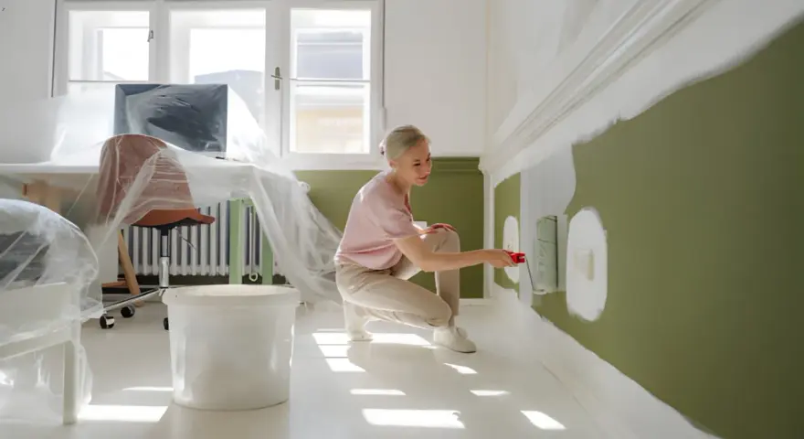 Kobieta maluje ścianę na zielono, klęcząc obok wiadra z farbą.
