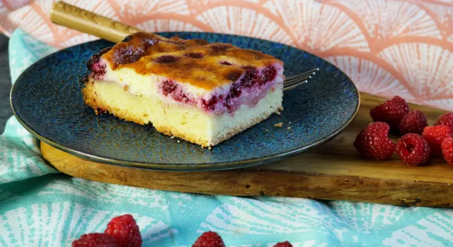 Ein Stück Himbeer Schmand Kuchen auf einem blauen Teller, daneben frische Himbeeren.