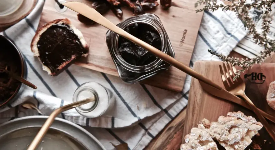 Słodkie śniadanie: kromka chleba z kremem czekoladowym, słoiczek z ciemnym dżemem, daktyle i ciastka. Idealna nutella bez orzechów!