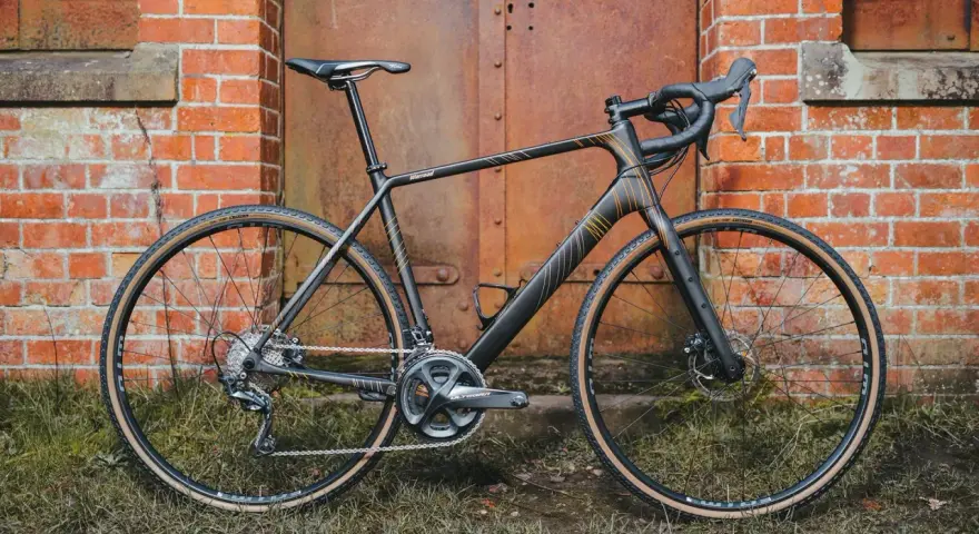 A sleek black Salsa Warroad gravel bike with orange accents stands against a rustic brick wall and weathered metal door.