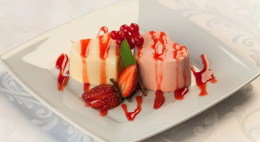 Dwie panna cotta w kształcie serc, polane sosem truskawkowym, ozdobione owocami i miętą.
