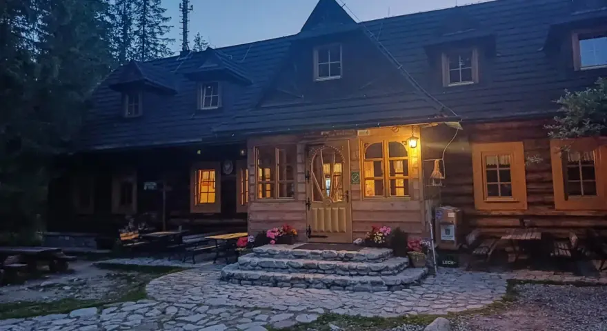 Drewniana chata w górach, oświetlona ciepłym światłem, to idealna baza pod roztokami. Kamienna ścieżka prowadzi do wejścia.