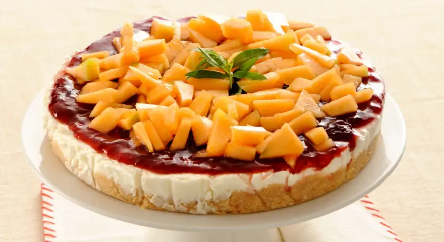Deliziosa cheesecake al melone, con topping di frutta fresca e salsa rossa, su un piatto bianco.