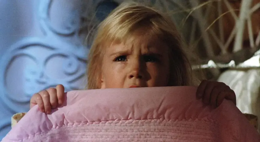 Bambina spaventata si nasconde sotto una coperta rosa, gli occhi sgranati in un'espressione di puro horror psicologico.