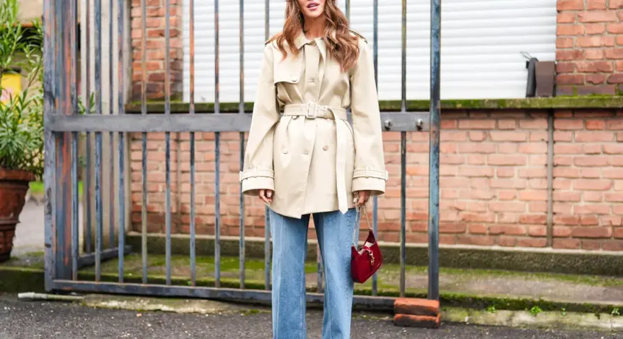 Mujer con gabardina beige y vaqueros bootcut, gafas rojas y bolso rojo.