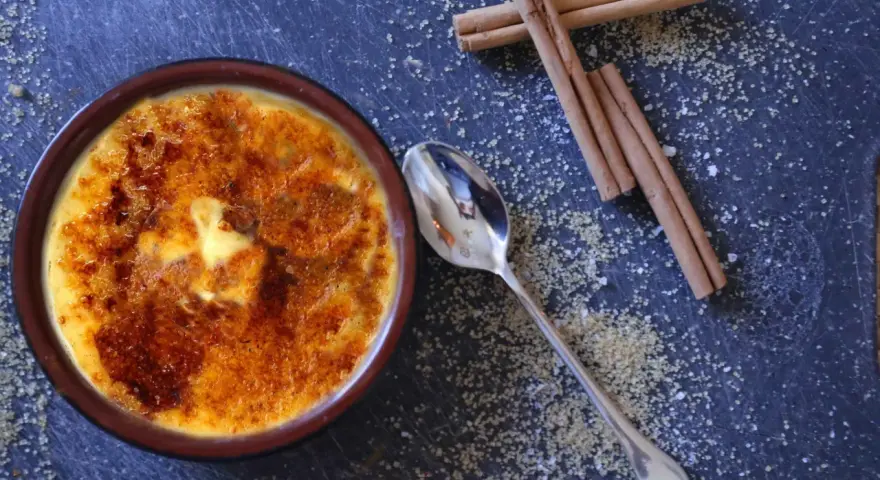 Cremige Crema Catalana mit karamellisierter Zuckerkruste, daneben Zimtstangen und Zucker.