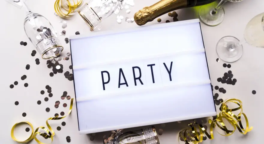 Świętowanie zaczyna się od zaproszeń! Na stole leżą kieliszki, szampan i konfetti, a tablica świetlna głosi "PARTY".