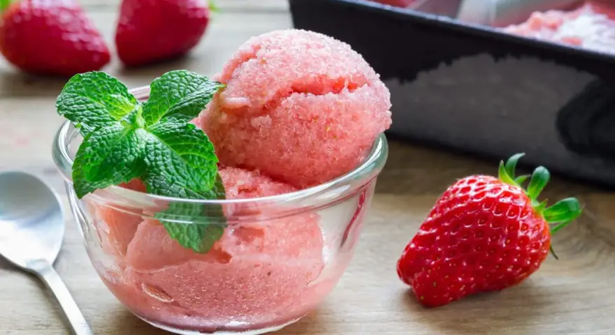Erfrischendes Erdbeer-Sorbet-Eis in einer Glasschale, garniert mit Minze. Daneben liegen frische Erdbeeren.