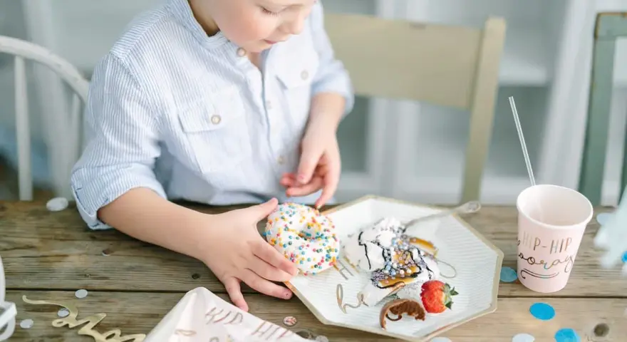 En pojke med en randig skjorta tar en donut med strössel. Det är en festlig dukning med konfetti och en mugg med texten "Hip Hip Hooray". En härlig stund av burken lek.
