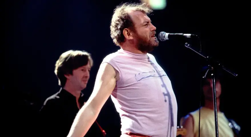 Joe Cocker śpiewa na scenie, a jego charakterystyczne utwory niosą się po sali. W tle widać innych muzyków.
