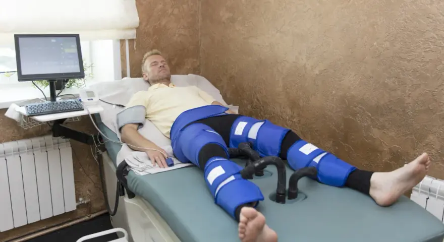 Mężczyzna leży na łóżku terapeutycznym, podłączony do aparatury do limfodrenażu. Widoczne są jego nogi w specjalnych mankietach, które mają zapewnić widoczne limfodrenaż efekty.
