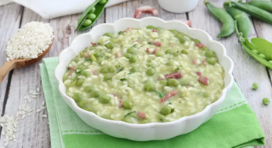Un cremoso piatto di risi e bisi, con piselli freschi e cubetti di pancetta, servito in una ciotola bianca su un tovagliolo verde.