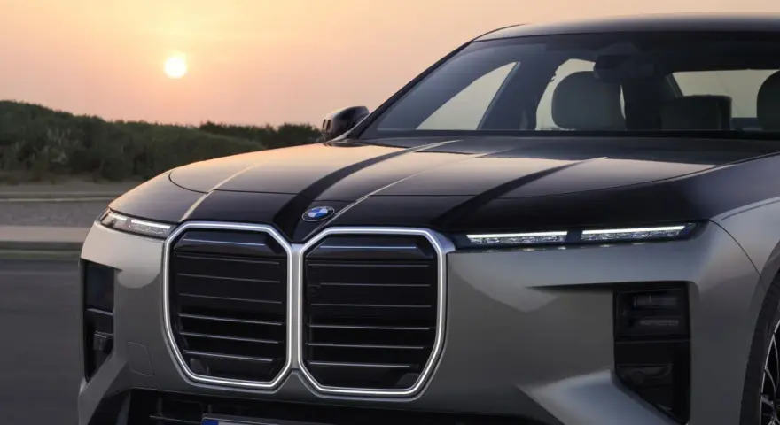 Przód luksusowego BMW M7 z podświetlaną atrapą chłodnicy i nowoczesnymi światłami, na tle zachodzącego słońca.
