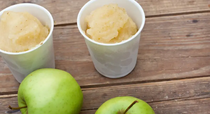Zwei Becher mit erfrischendem Apfel Sorbet stehen neben zwei grünen Äpfeln auf einer Holzoberfläche.