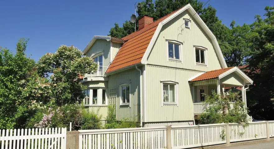 Ljusgrönt trähus med tegeltak och vit staket. En vacker trädgård med blommande buskar. Planerar att byta fasad för att fräscha upp huset.