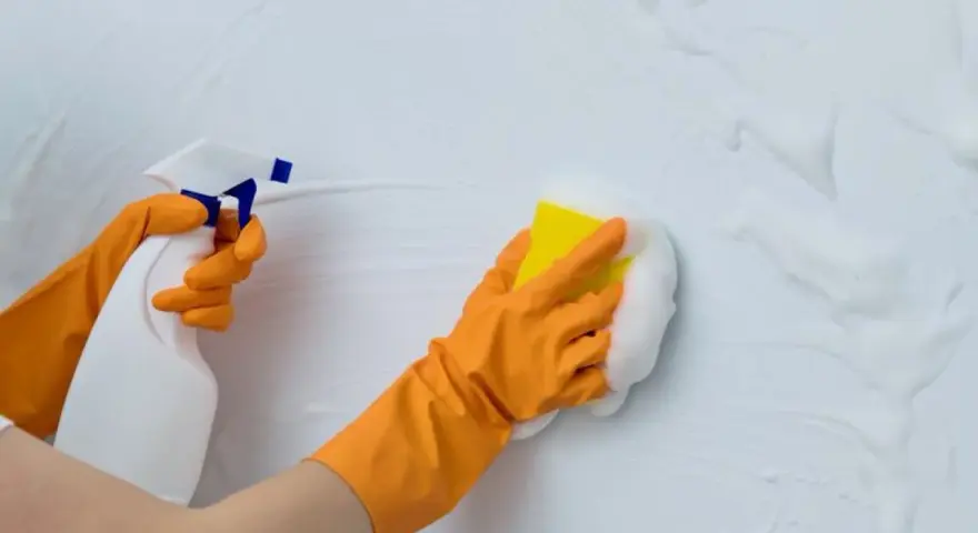 Manos con guantes naranjas rociando y limpiando paredes con espuma y esponja.