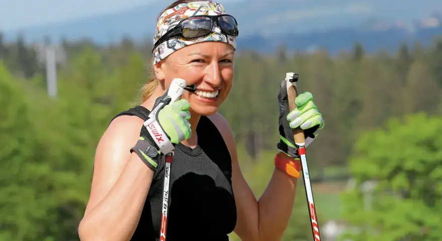 Uśmiechnięta Justyna Kowalczyk-Tekieli z kijkami do nartorolkarstwa, w sportowym stroju i opasce na głowie, gotowa do treningu.