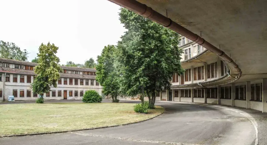 Alte Gebäude im olympisches Dorf Berlin, umgeben von Bäumen und Grünflächen. Eine Straße führt an einem geschwungenen Gebäude mit vielen Fenstern vorbei.
