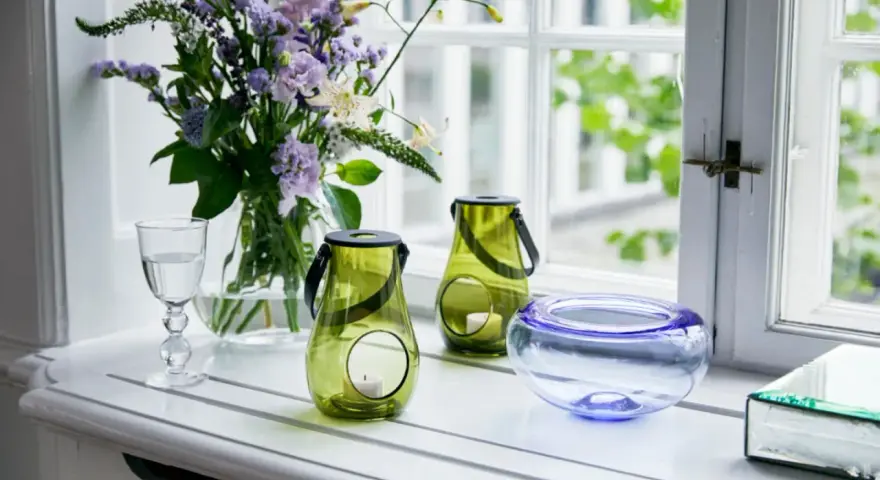 Ein Strauß lila Blumen und grüne Windlichter schmücken die Fensterbank. Ein Glas Wasser und eine blaue Schale runden die Dekoration ab.