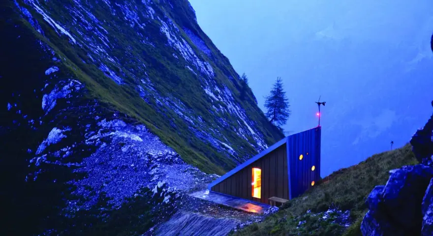 Un moderno bivacco interno, illuminato da una luce calda, si staglia su un pendio montuoso al crepuscolo.