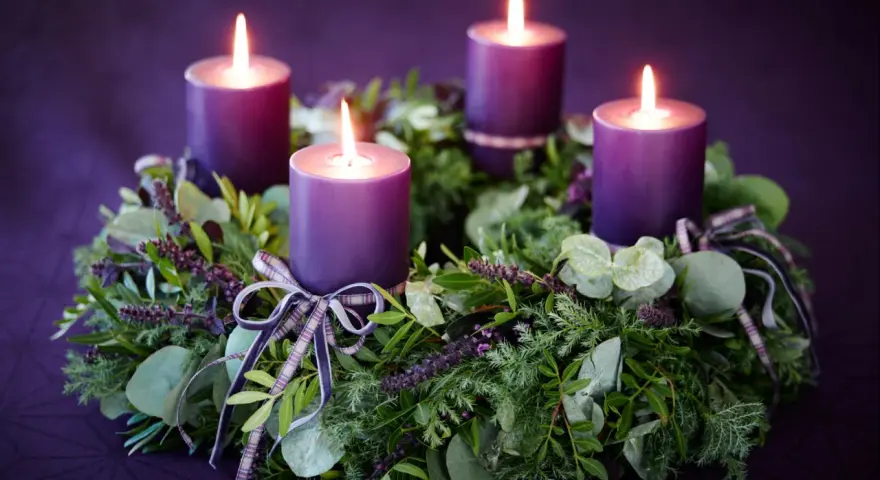 Four purple candles glow on a lush green Advent wreath, adorned with ribbons. The final candle's flame flickers, marking the 4th Advent.