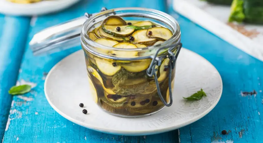 Barattolo di zucchine in agrodolce, con pepe nero e foglie di menta, su un piatto bianco appoggiato su un tavolo blu.