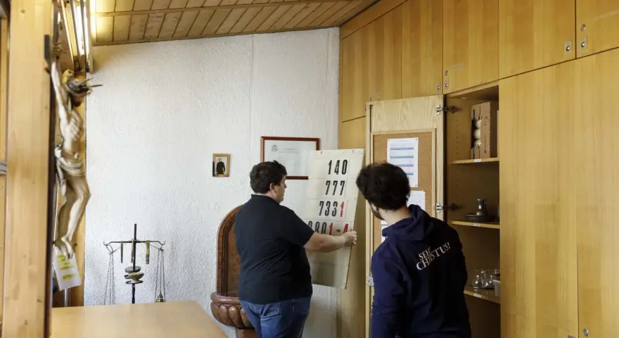 Zwei Männer im katholischen Priesterseminar hängen eine Liste mit Zahlen auf. Ein Kruzifix hängt an der Wand.