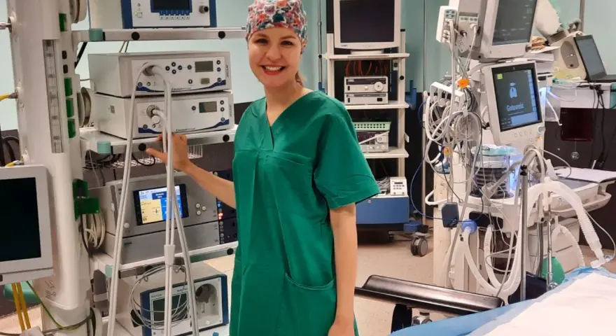 Uśmiechnięta pielęgniarka operacyjna, ubrana w zielony fartuch i czepkek, stoi obok stosu aparatury medycznej. Obok niej widać instrumentariuszka przygotowująca się do zabiegu.