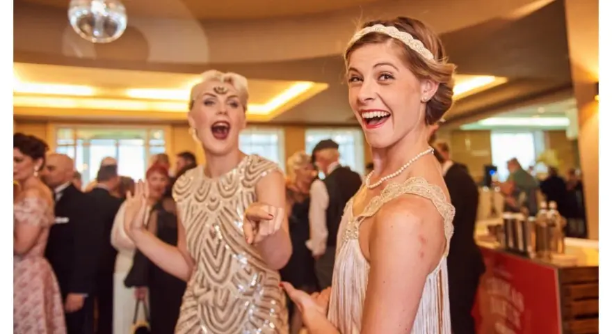 Deux femmes dansent, l'une porte une robe à paillettes dorées et un bandeau, l'autre une robe à franges, parfaites pour une tenue guinguette femme.