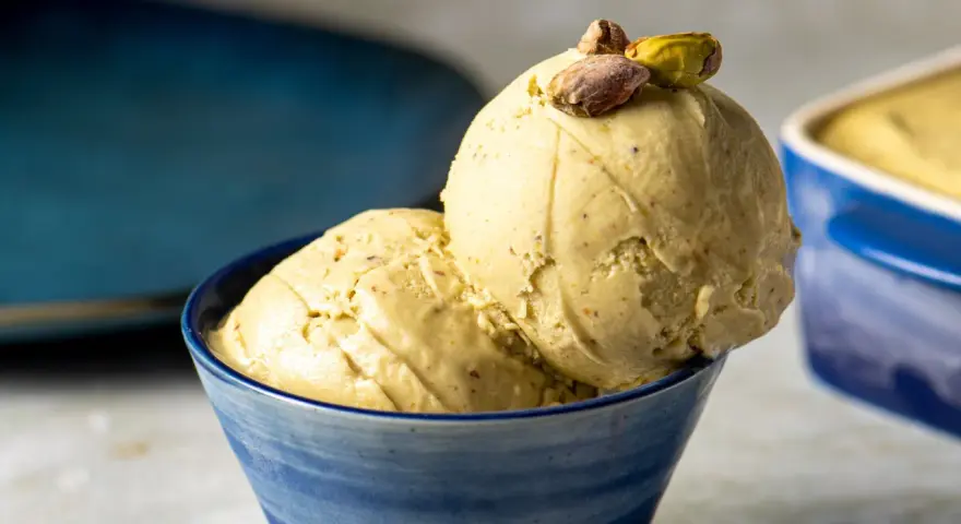 Zwei Kugeln cremiges, gelbes Eis mit Pistazienstückchen, garniert mit ganzen Pistazien, in einer blauen Schale.