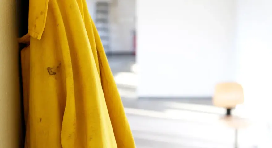 Gelbe Regenjacke hängt an einer Wand, bereit für die Regenjacke reparieren. Flecken auf dem Stoff deuten auf vergangene Abenteuer hin.