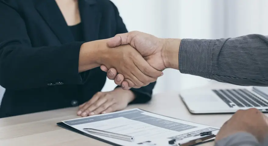 Dos personas se dan la mano sobre un escritorio con un currículum y un portátil. Es el final de una entrevista en francés.