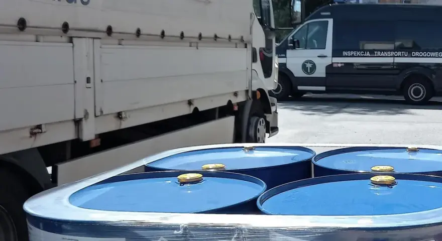 Niebieskie beczki z żółtymi zamknięciami, przygotowane do transportu towarów niebezpiecznych, obok ciężarówki i radiowozu Inspekcji Transportu Drogowego.