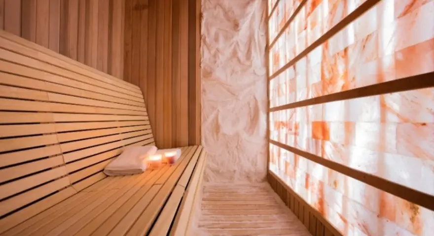 A serene salt sauna with a wooden bench, towels, candles, and a wall of glowing Himalayan salt bricks.