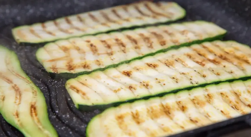 Fette di zucchine grigliate che cuociono su una piastra, con segni di grigliatura dorati e invitanti.
