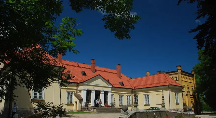 Pałac w Waplewie Wielkim, żółty, z czerwonym dachem, otoczony zielenią, pod błękitnym niebem.