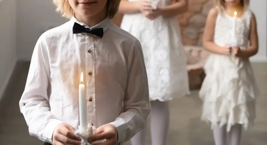 Dziecko w białej koszuli z czarną muszką trzyma zapaloną świecę. W tle inne dzieci w białych strojach.