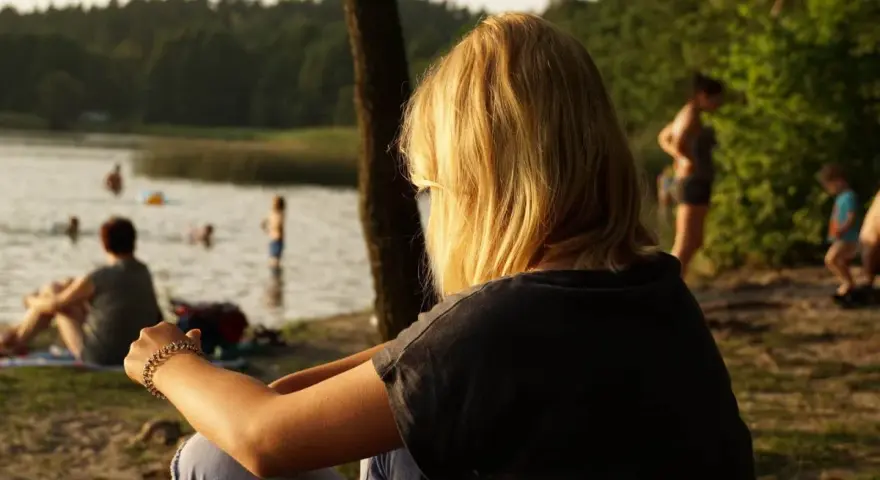 Dziewczyna z blond włosami siedzi nad jeziorem Trzechowskim, obserwując ludzi kąpiących się w wodzie.