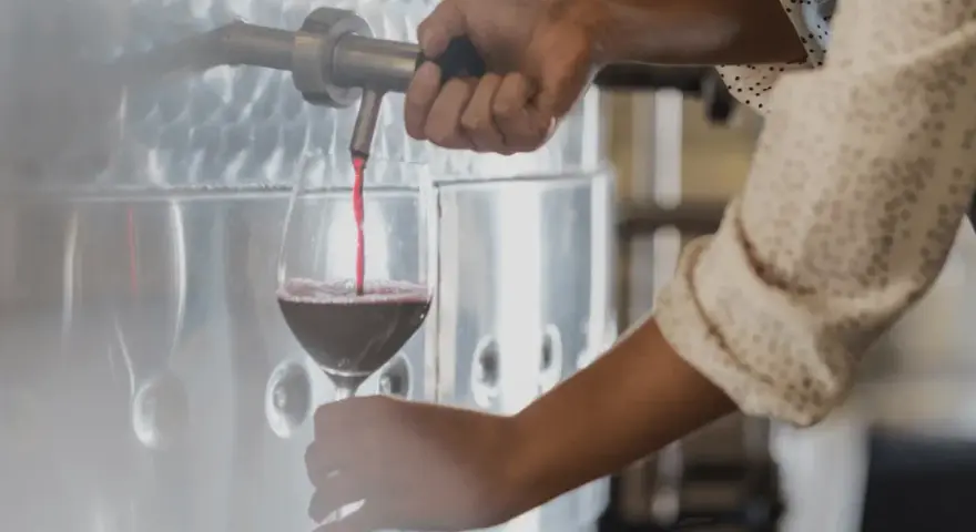 Rött vin hälls upp i ett glas från en stor tank. En hand vrider på kranen, kanske för att kontrollera tillsatser i vin.