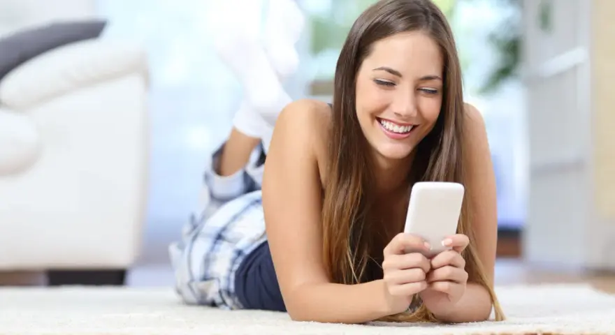Uśmiechnięta dziewczyna w piżamie leży na dywanie, przeglądając telefon. Może szuka nowych znajomości na Tinder randki.