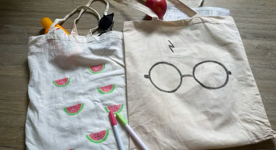 Zwei jute beutel bemalen: einer mit Wassermelonen, der andere mit Harry Potters Brille. Sonnencreme, Sonnenbrille und Stifte liegen daneben.