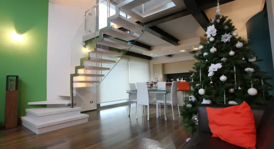 Un moderno soppalco casa con scala in vetro e legno, albero di Natale addobbato e un divano con cuscino arancione.