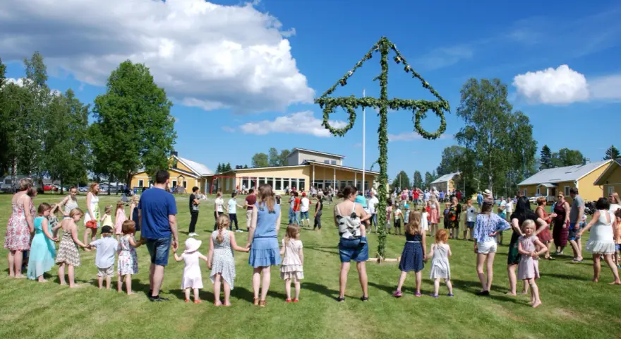 Många människor, både vuxna och barn, håller varandra i händerna och dansar runt en midsommarstång. De firar midsommar i en grönskande park med gula byggnader i bakgrunden.