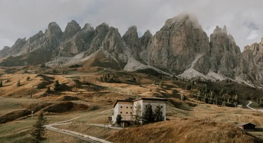 Malowniczy krajobraz w dolinie Val di Funes z budynkiem hotelowym na wzgórzu i skalistymi szczytami w tle.