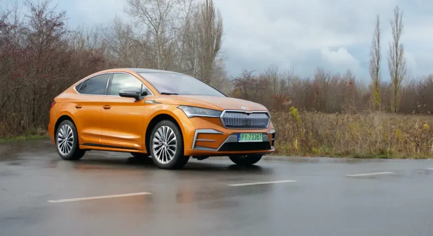 Pomarańczowa Skoda Enyaq Coupe na mokrej drodze, zarośla w tle.