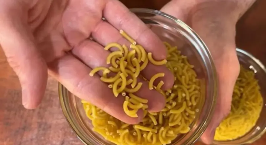 Mano sosteniendo fideos cortos y curvos sobre un bol de fideos similares.