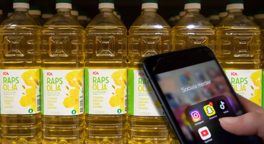 En hand håller upp en mobil med sociala medier-appar. Bakom syns rader av rapsoljeflaskor, där en del anser att rapsolja är farligt.