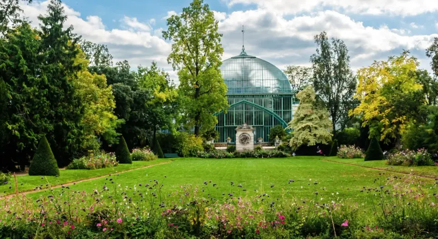 Un jardín exuberante en el Bois de Boulogne, con una gran estructura de cristal verde y un cielo azul con nubes.